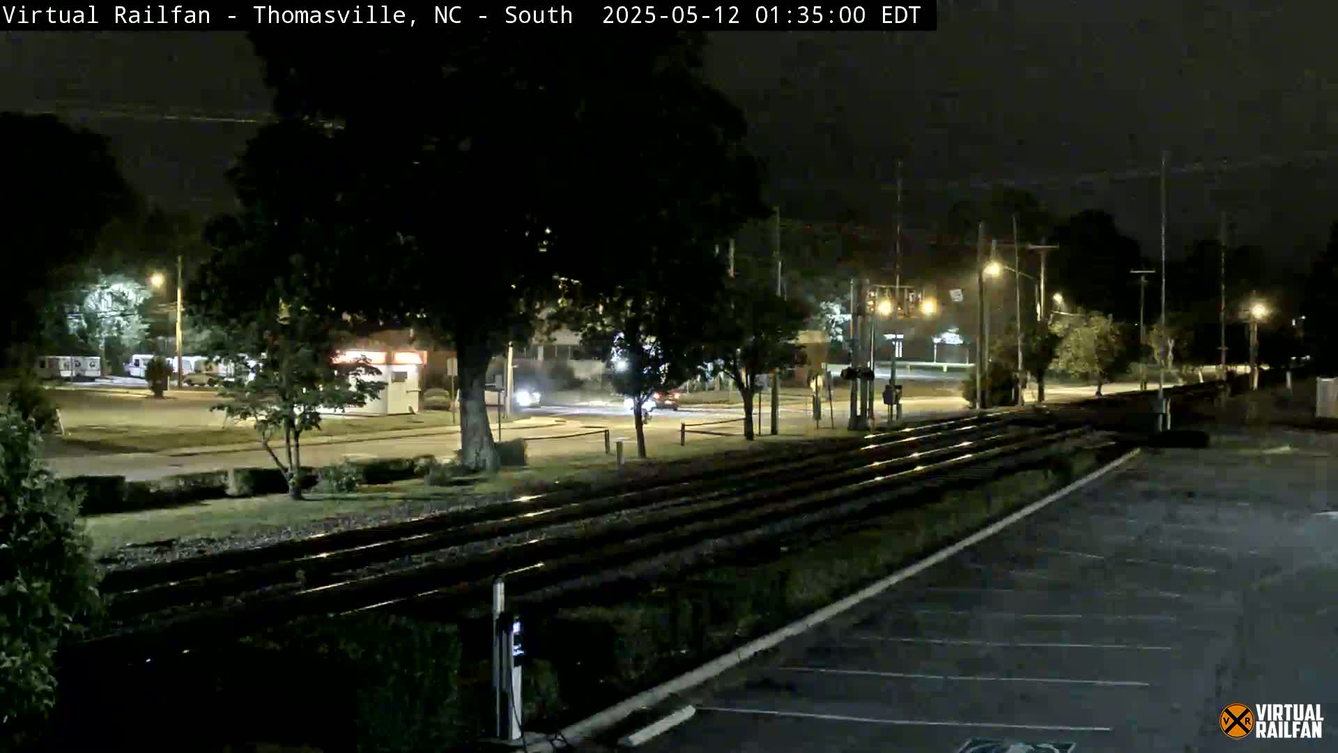 At night, a view of a street and train tracks shows several vehicles driving on the road next to a parking lot under streetlights.