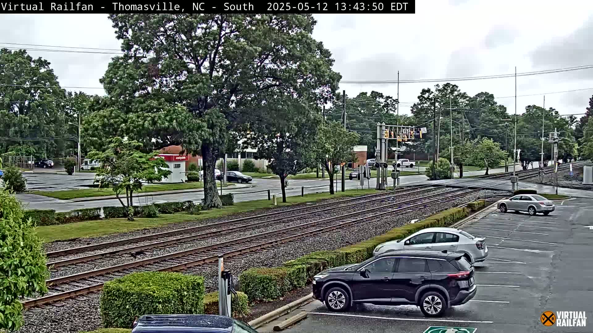 A view of train tracks and a street intersection on an overcast day, with several cars parked in a lot alongside the tracks and a few more driving on the road.