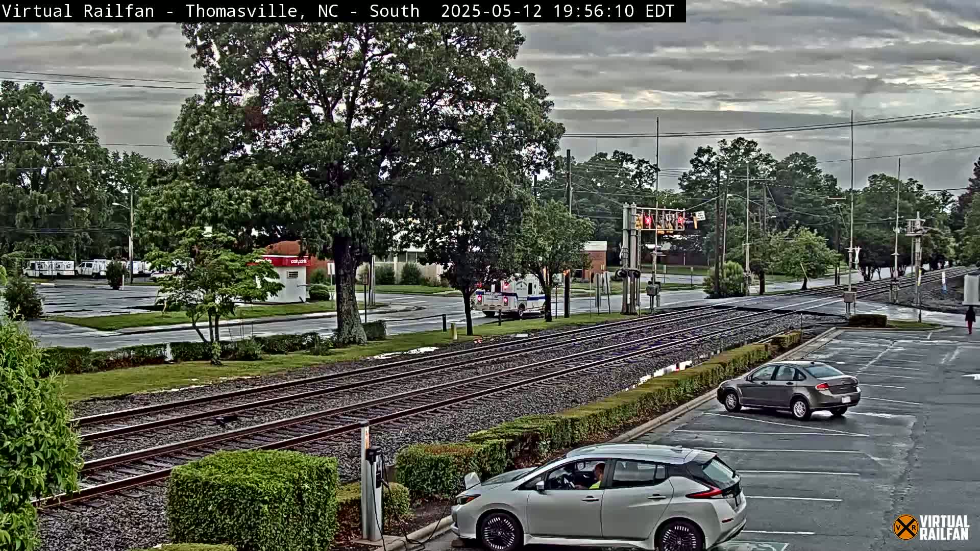 A view of railroad tracks intersecting a road, with several cars parked nearby, under an overcast sky.