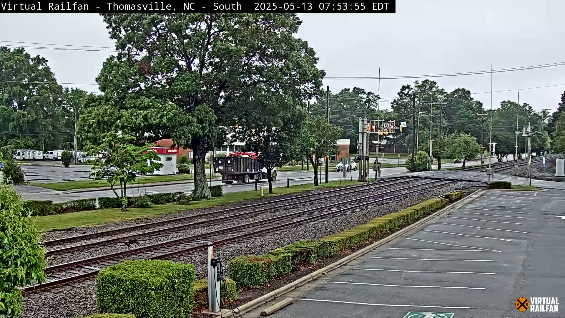 A dump truck is stopped at a railroad crossing on an overcast day, with several sets of train tracks running through a residential area lined with trees and shrubs.