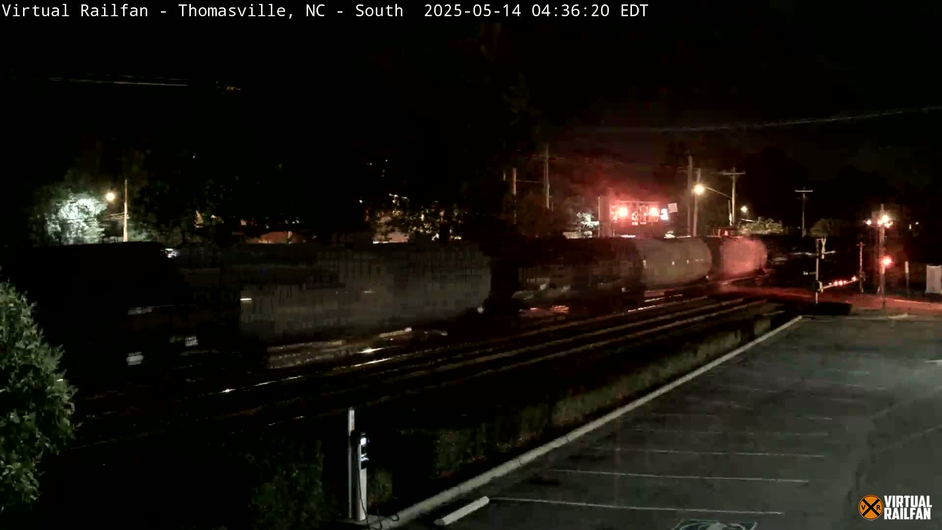 At night, a long freight train passes a railway crossing near a parking lot under dark, wet conditions.