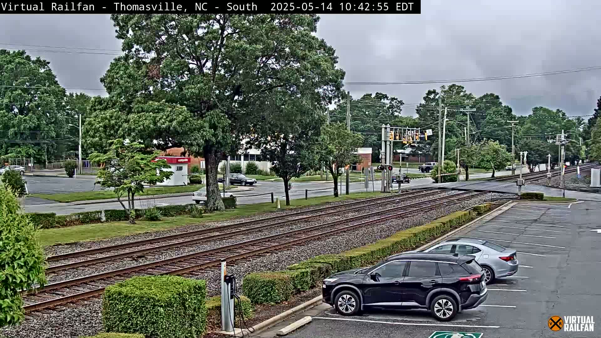 Train tracks intersect a road in a town under an overcast sky, with several cars parked nearby.