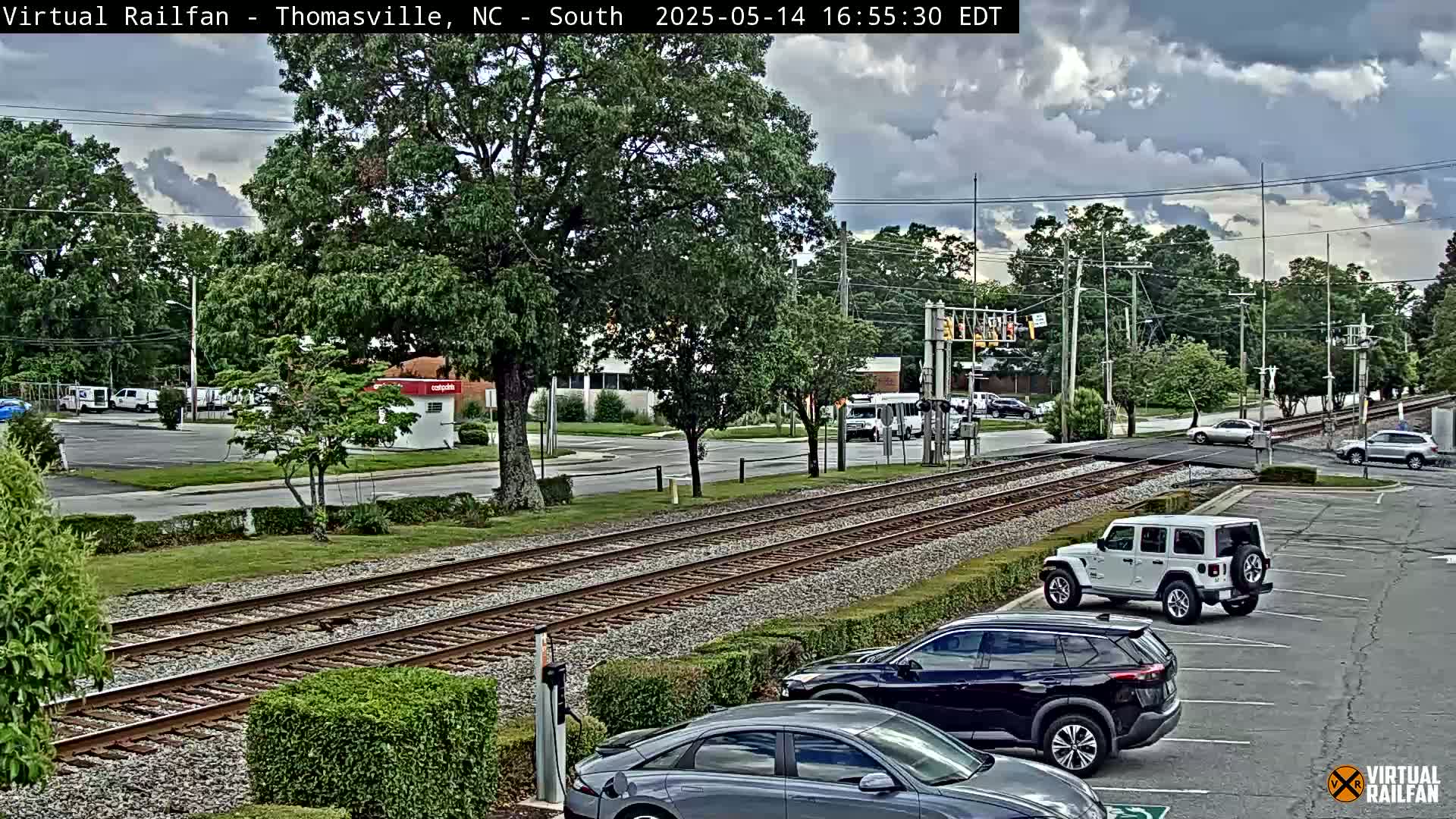 A parking lot with several cars is next to train tracks that cross a road under a partly cloudy sky.