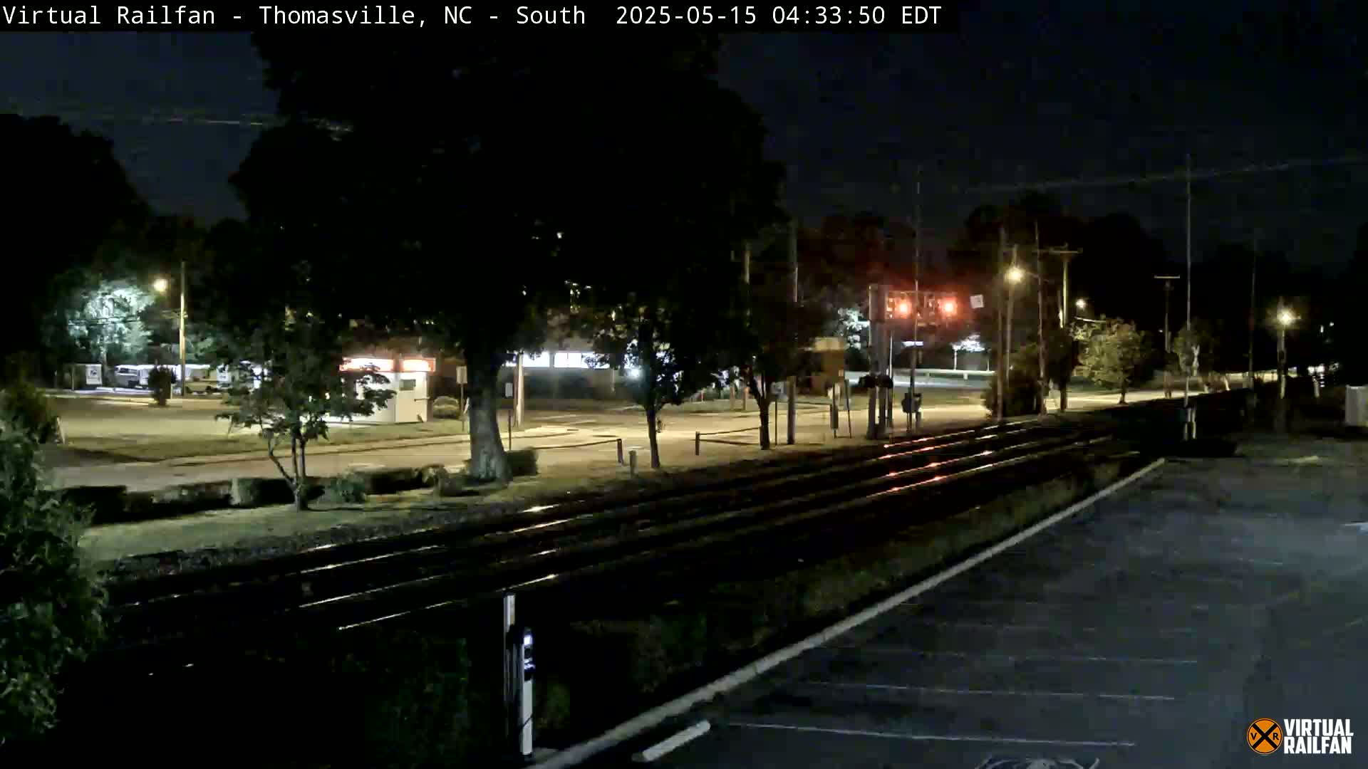 At night, a set of train tracks runs through a mostly empty parking lot adjacent to a road lined with trees and buildings under a clear, dark sky.