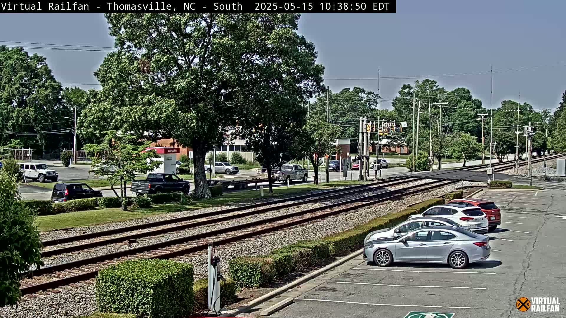 Under a clear sky, a train track runs through a town with cars parked in a lot and driving on the road beside it, surrounded by lush green trees and landscaping.