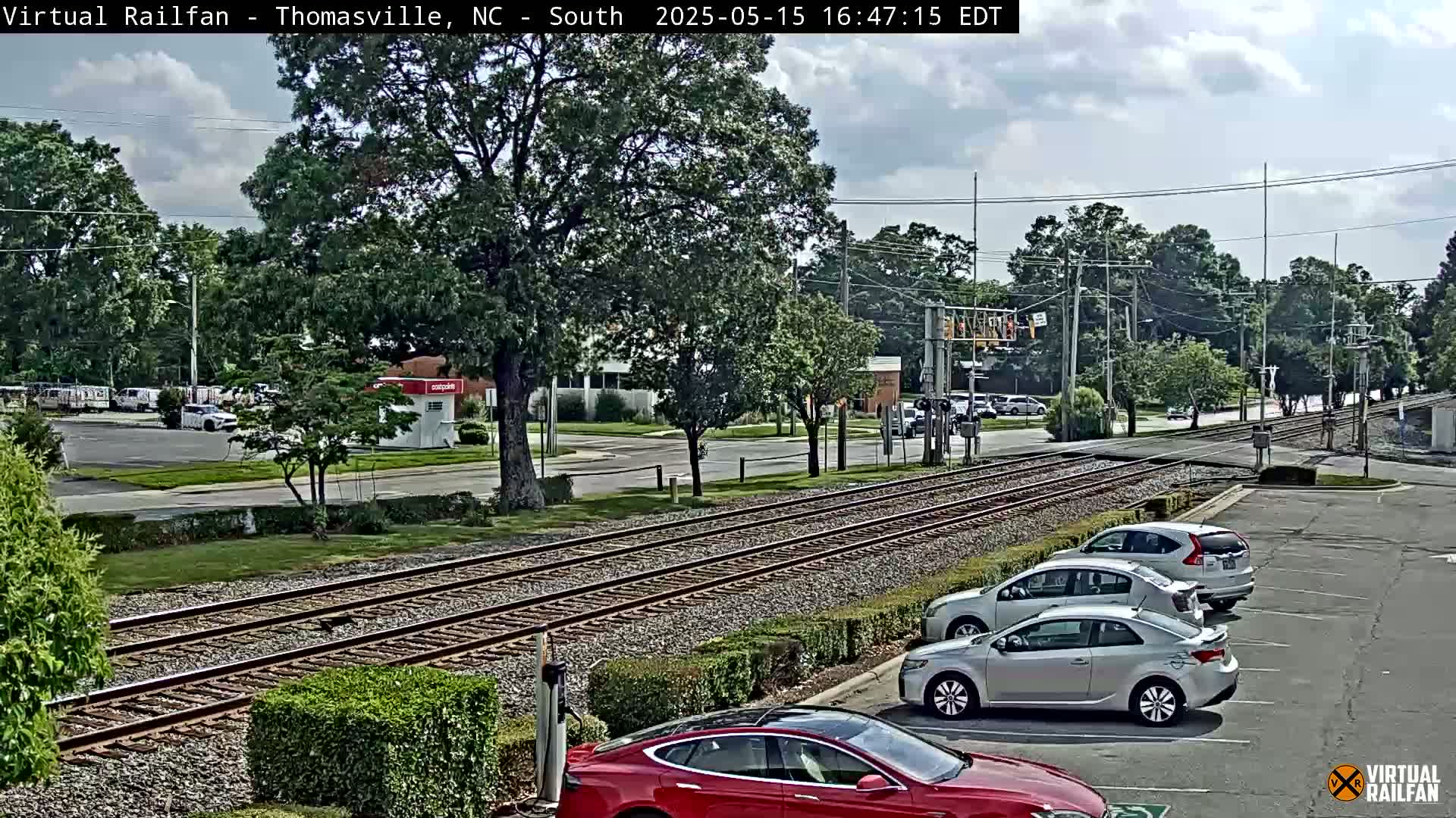 A parking lot with several cars is next to railroad tracks that cross a road under a partly cloudy sky.