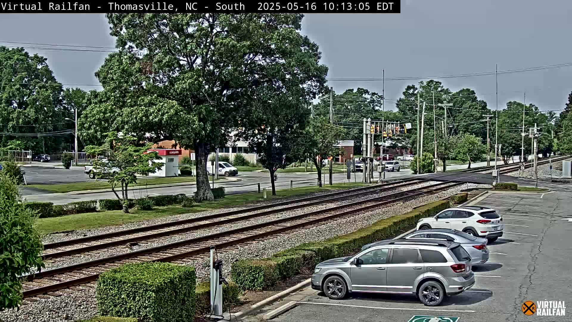 A sunny day shows railroad tracks running alongside a road with cars parked in a lot and driving on the road, with leafy trees and buildings in the background.