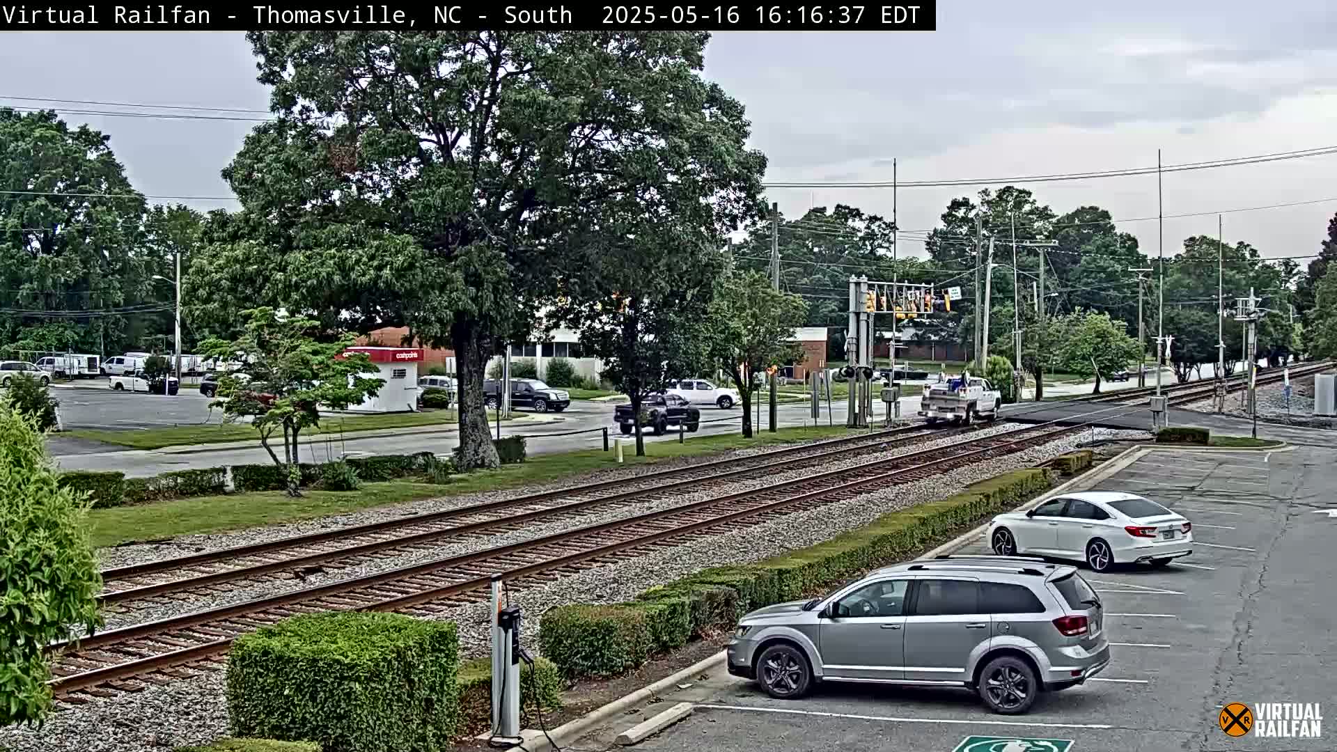 A mostly cloudy day shows a view of train tracks intersecting a road with several cars in a parking lot and at a nearby intersection.