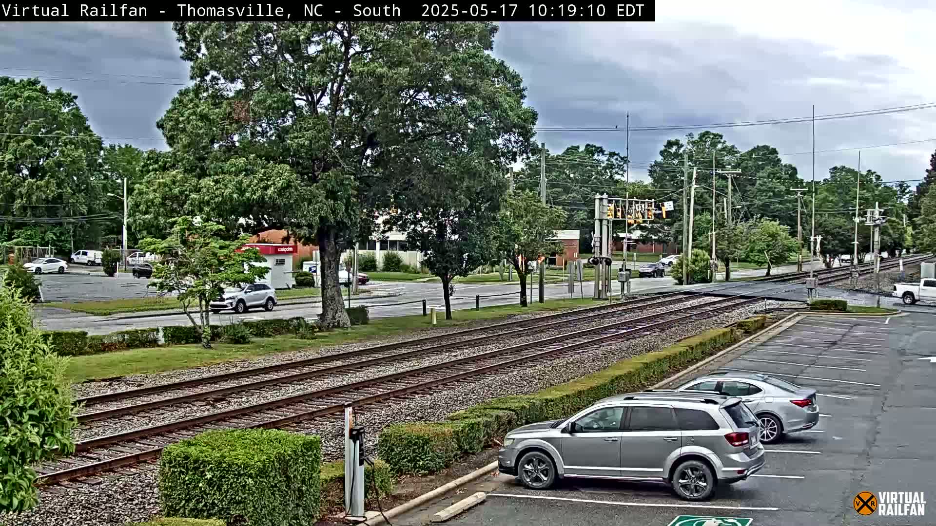 A view of railroad tracks intersecting a road, with several cars parked in a lot nearby, under an overcast sky.