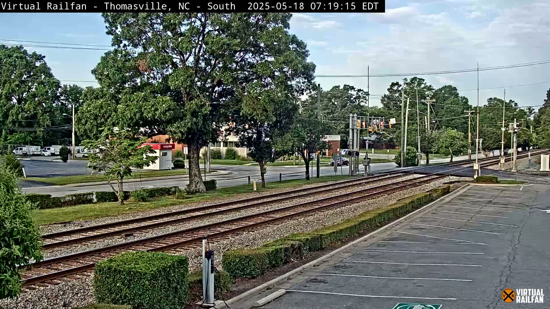 A mostly sunny view of railroad tracks crossing a street, surrounded by lush green trees and bushes, with a parking lot in the foreground.