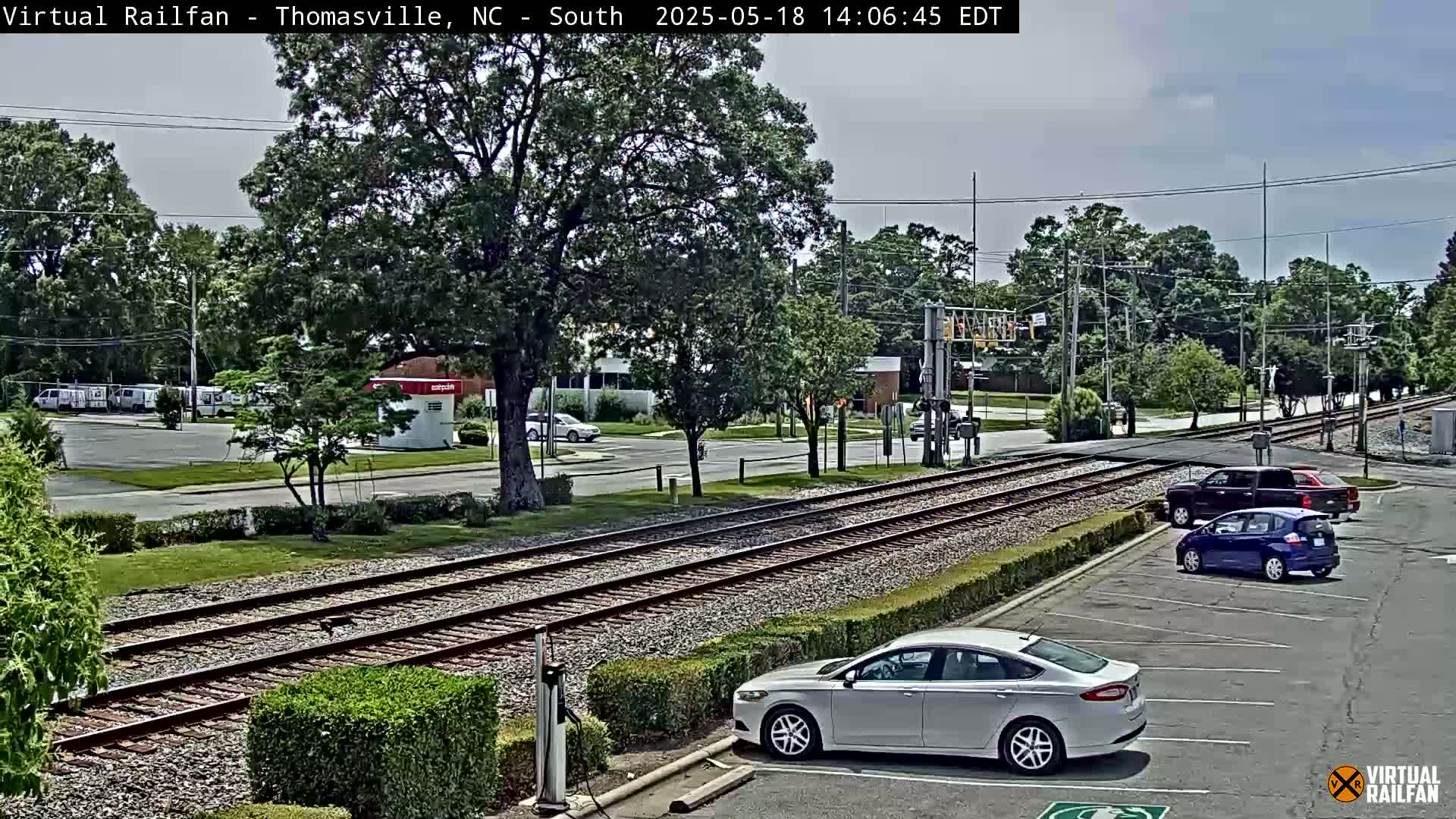 A mostly sunny view of train tracks crossing a road, with several cars parked nearby and lush green trees in the background.