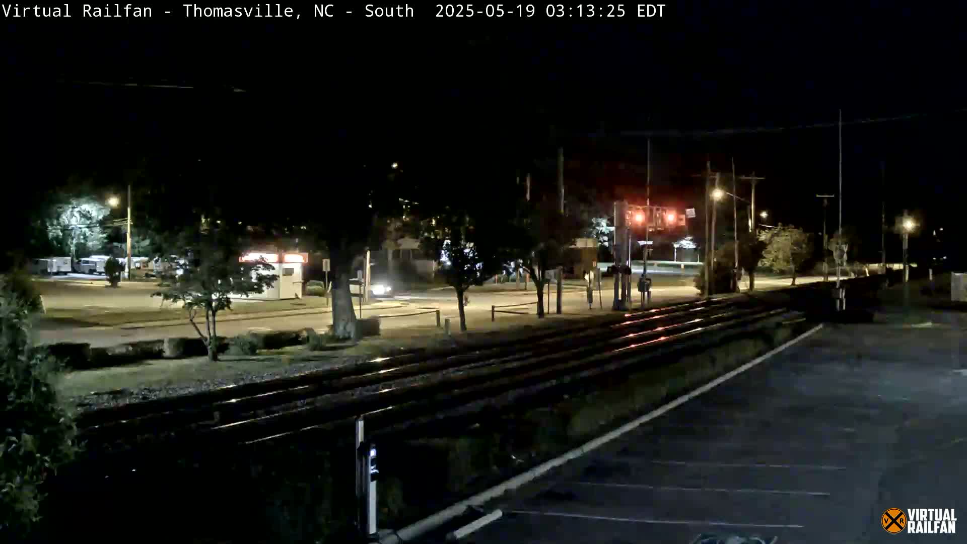 At night, a nighttime view shows train tracks next to a road with cars parked, and a few vehicles driving by under streetlights.