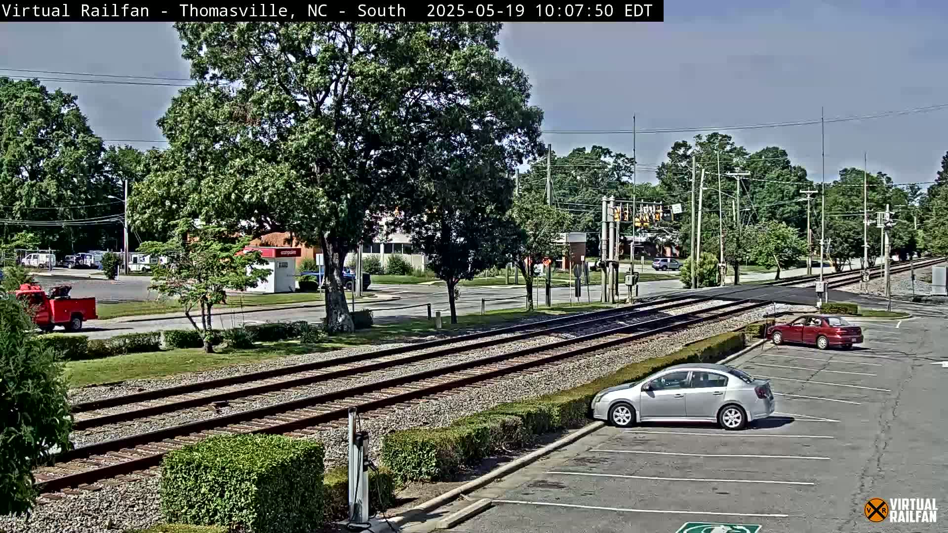 A sunny day shows railroad tracks crossing a road in a suburban area, with several cars parked and driving by.