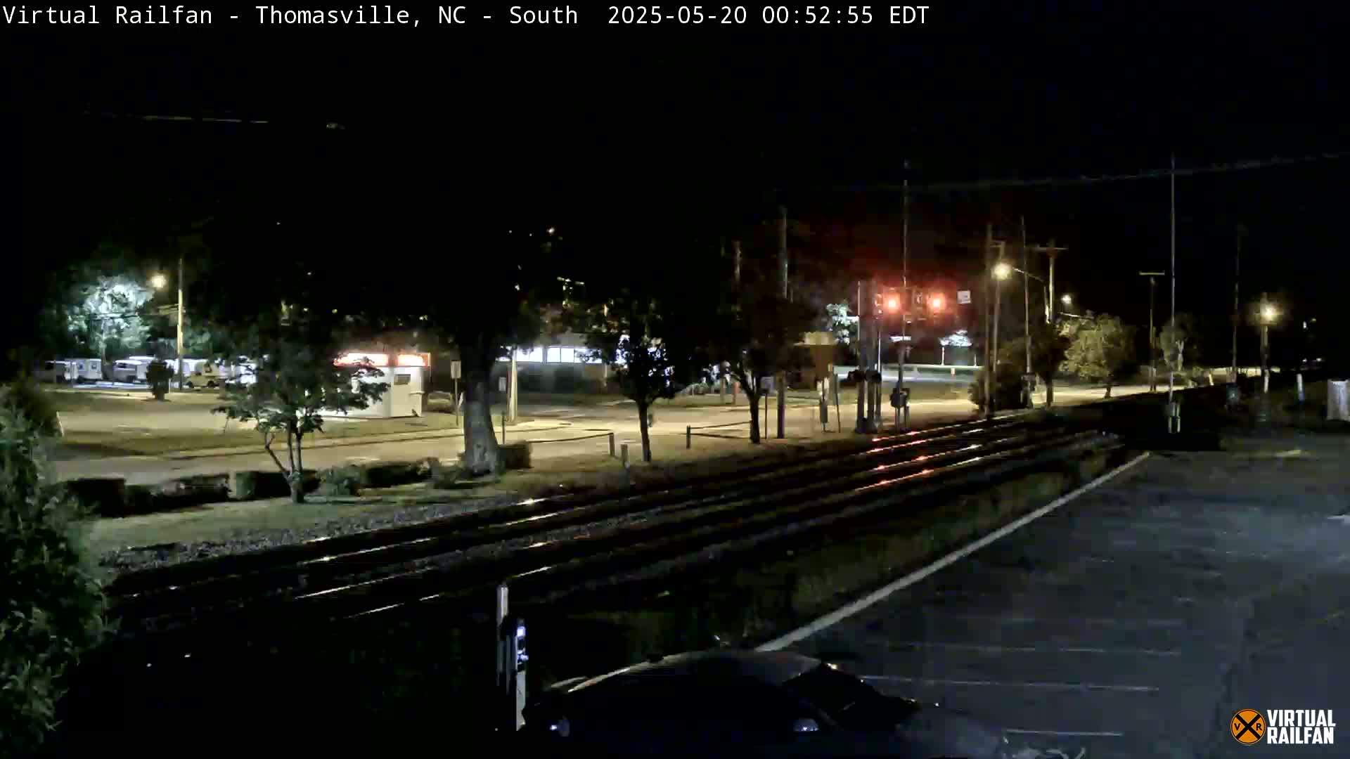 A nighttime view of railroad tracks next to a road, with some buildings and trees visible under streetlights.