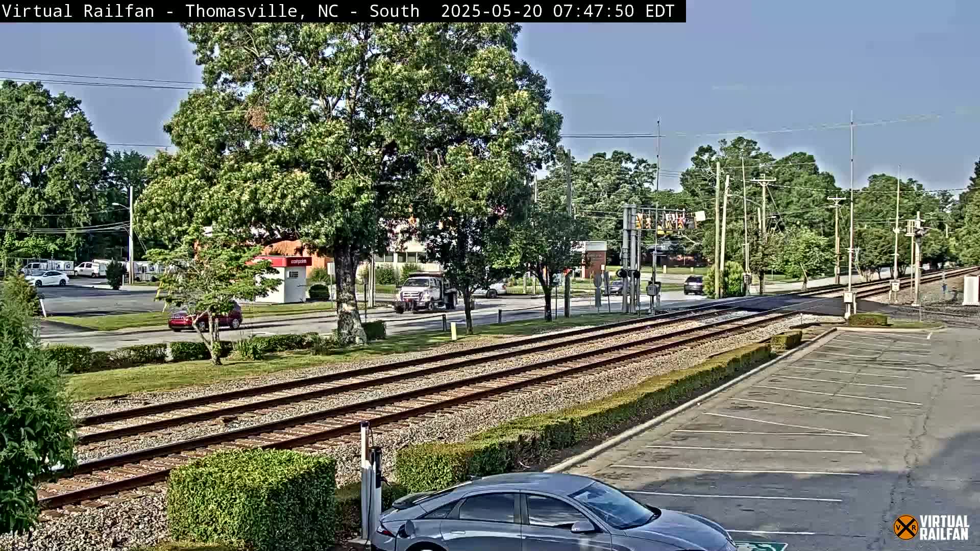 A sunny view of railroad tracks beside a road, with several cars visible, and lush green trees lining the streets.
