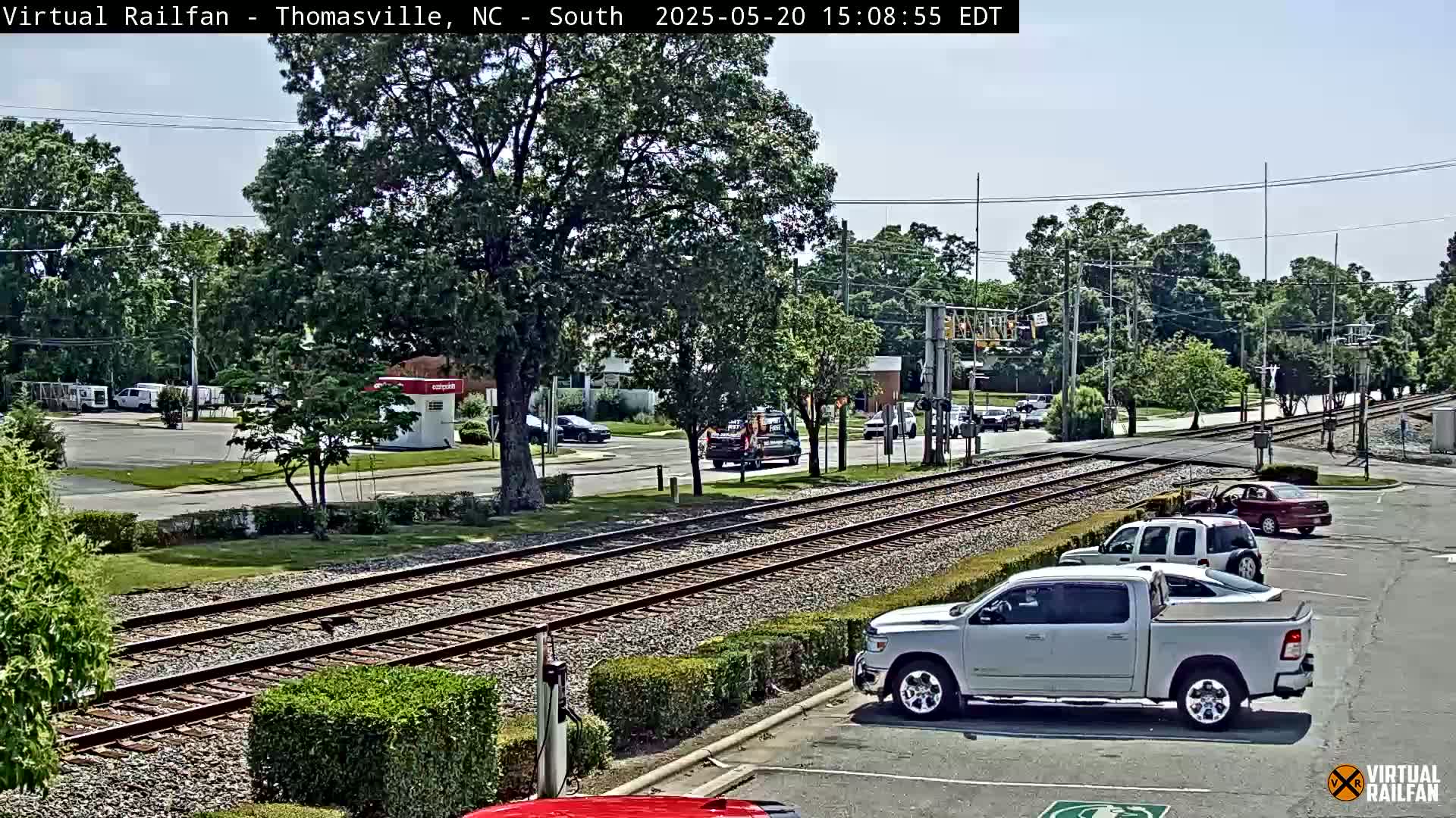 A sunny day view of railroad tracks running through a residential area with several vehicles parked and driving on adjacent roads and in a parking lot.