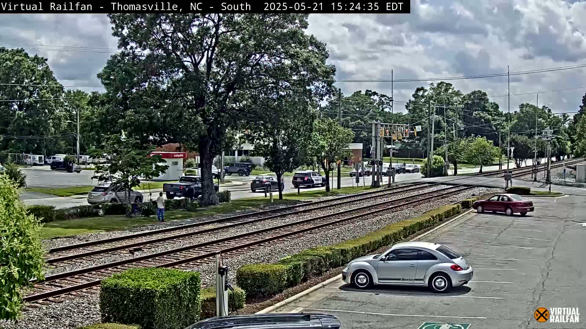 A partly cloudy day shows several cars on a road crossing railroad tracks, with a few people standing nearby.