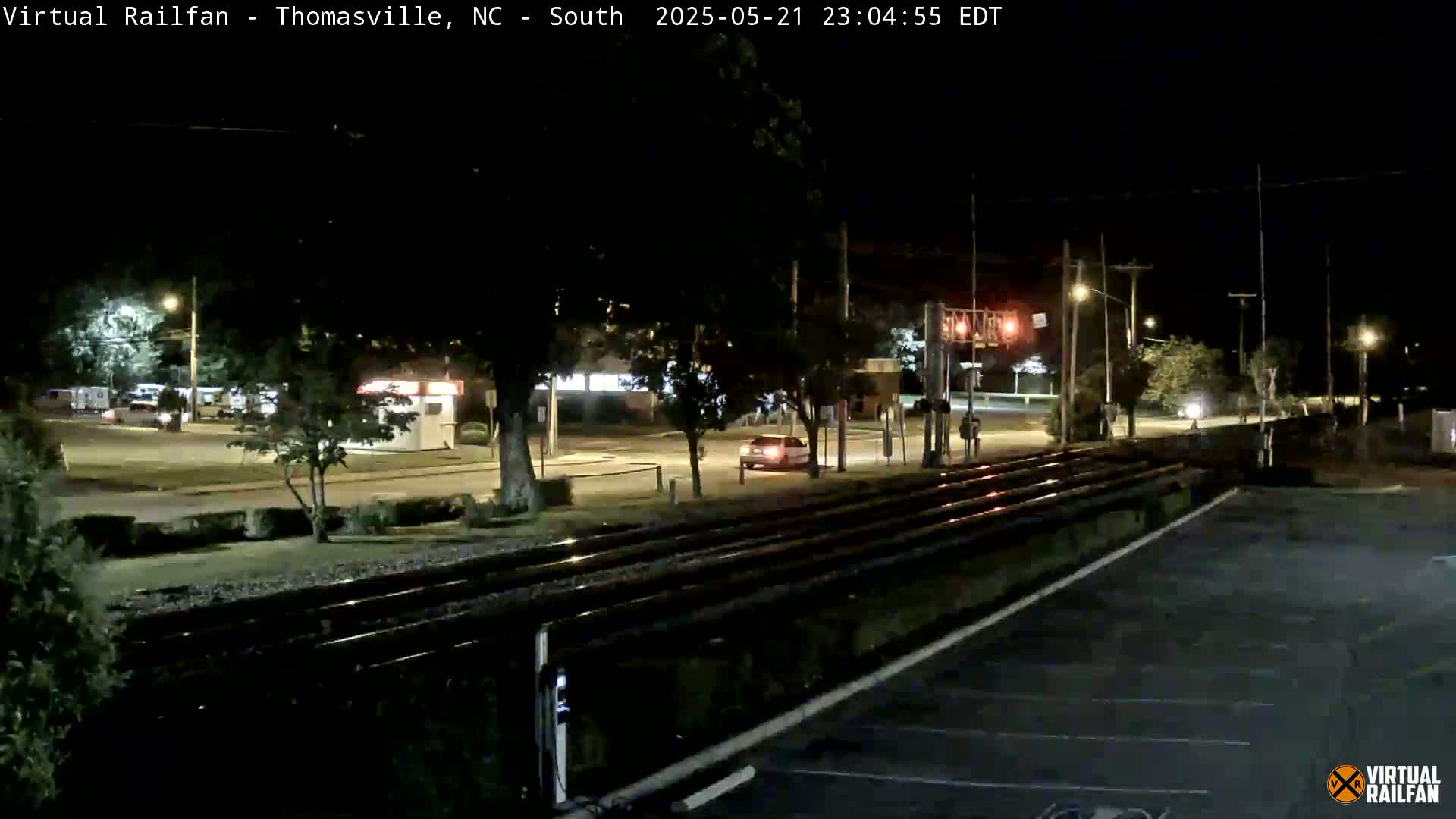 A nighttime view of a street intersection with a vehicle, railroad tracks, and streetlights, under clear skies.