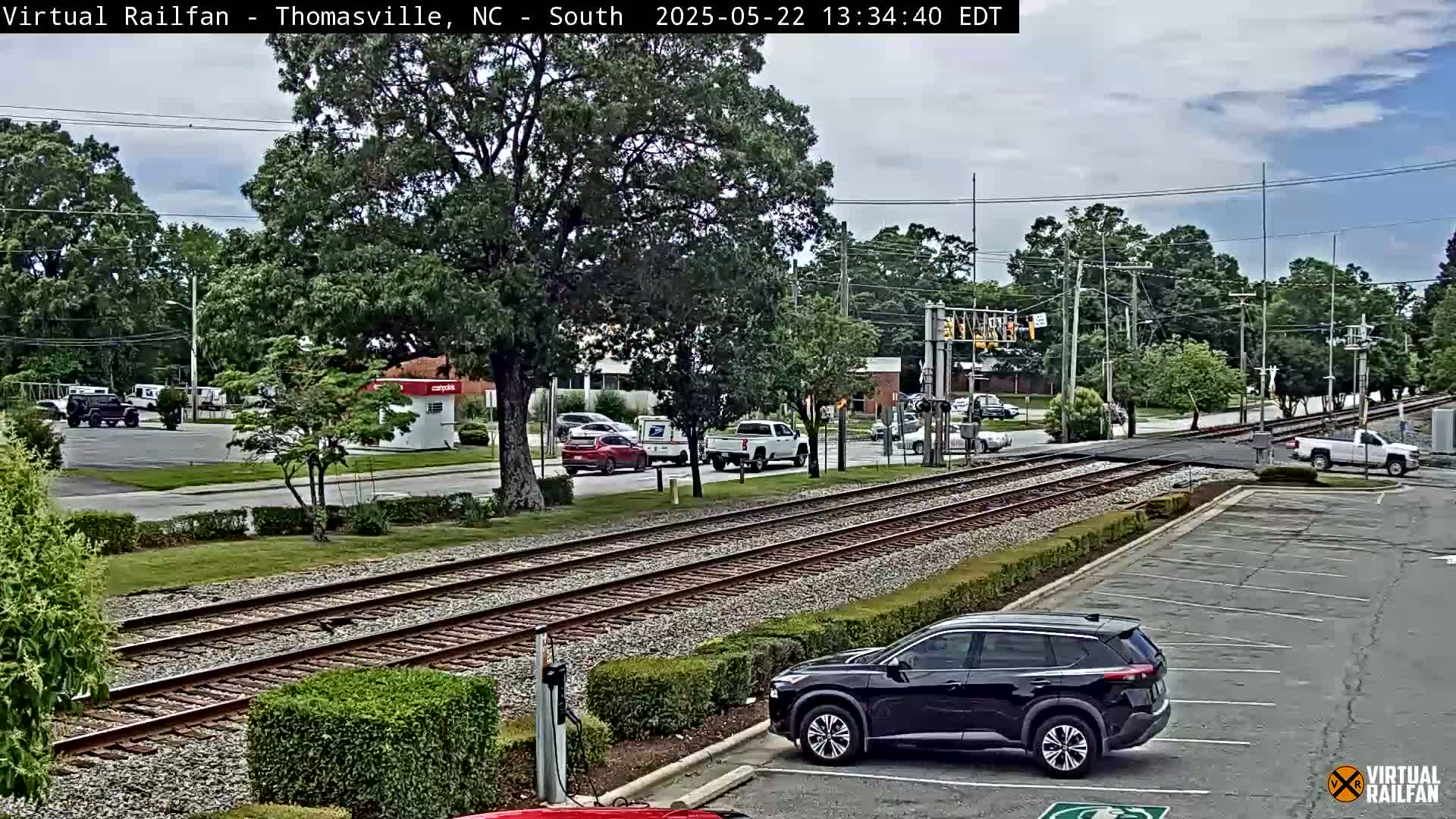 A mostly sunny day shows a train track intersecting a road with several vehicles parked and driving in a suburban area with lush green trees.
