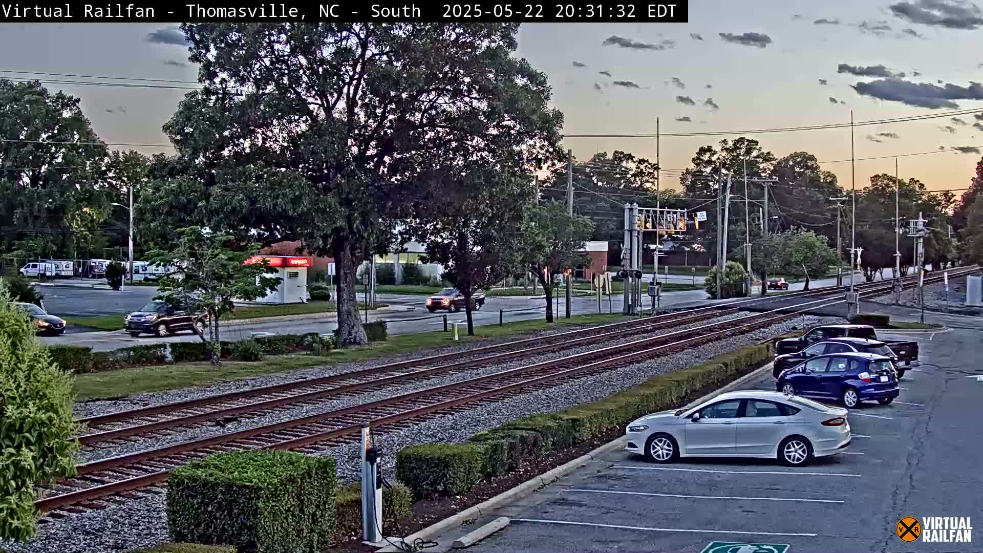 A twilight view of a railroad crossing with several cars parked and driving near a roadway, under a partly cloudy sky.
