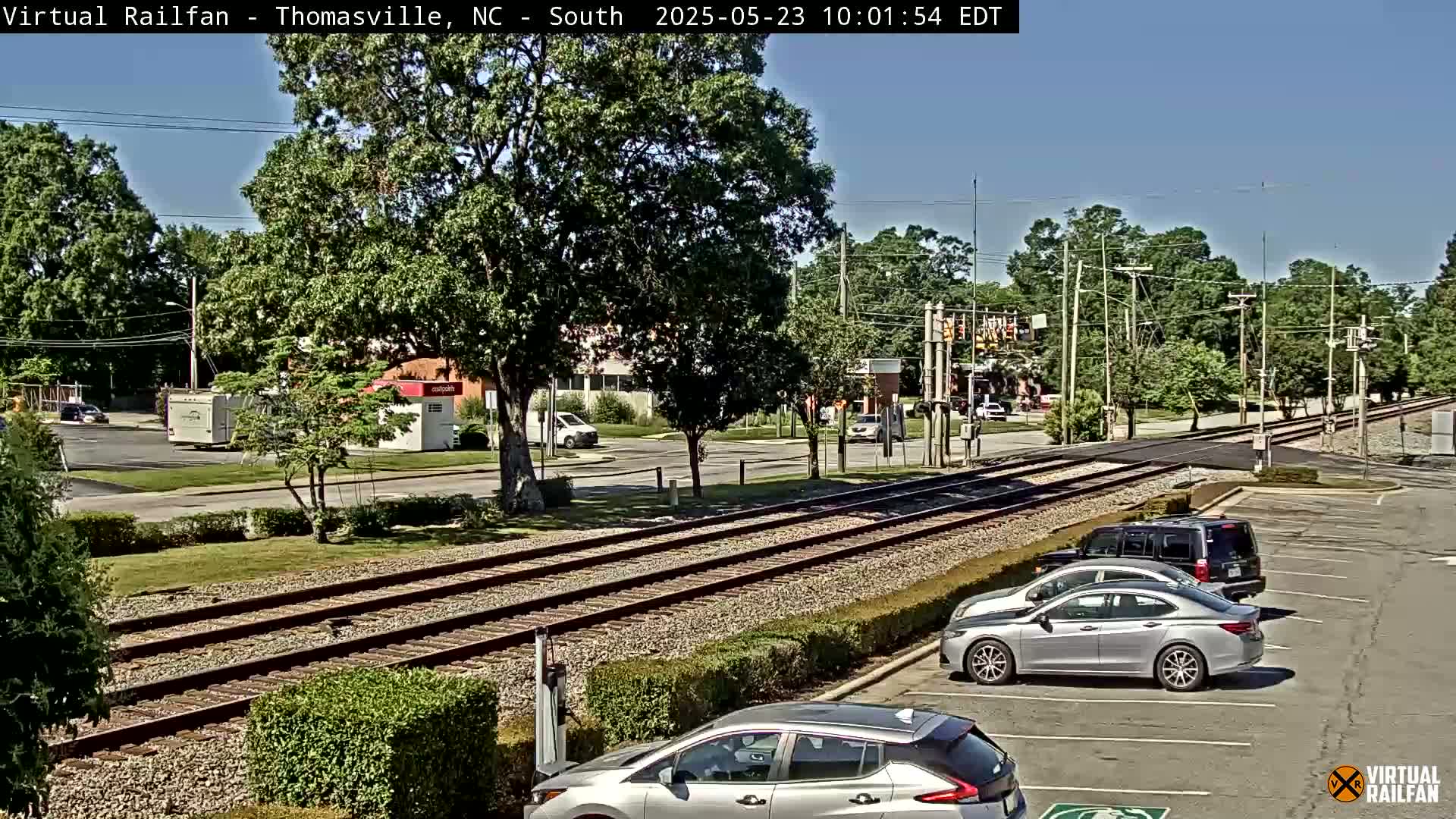A sunny day scene shows railroad tracks running through a residential area with cars parked nearby, and trees lining the streets.