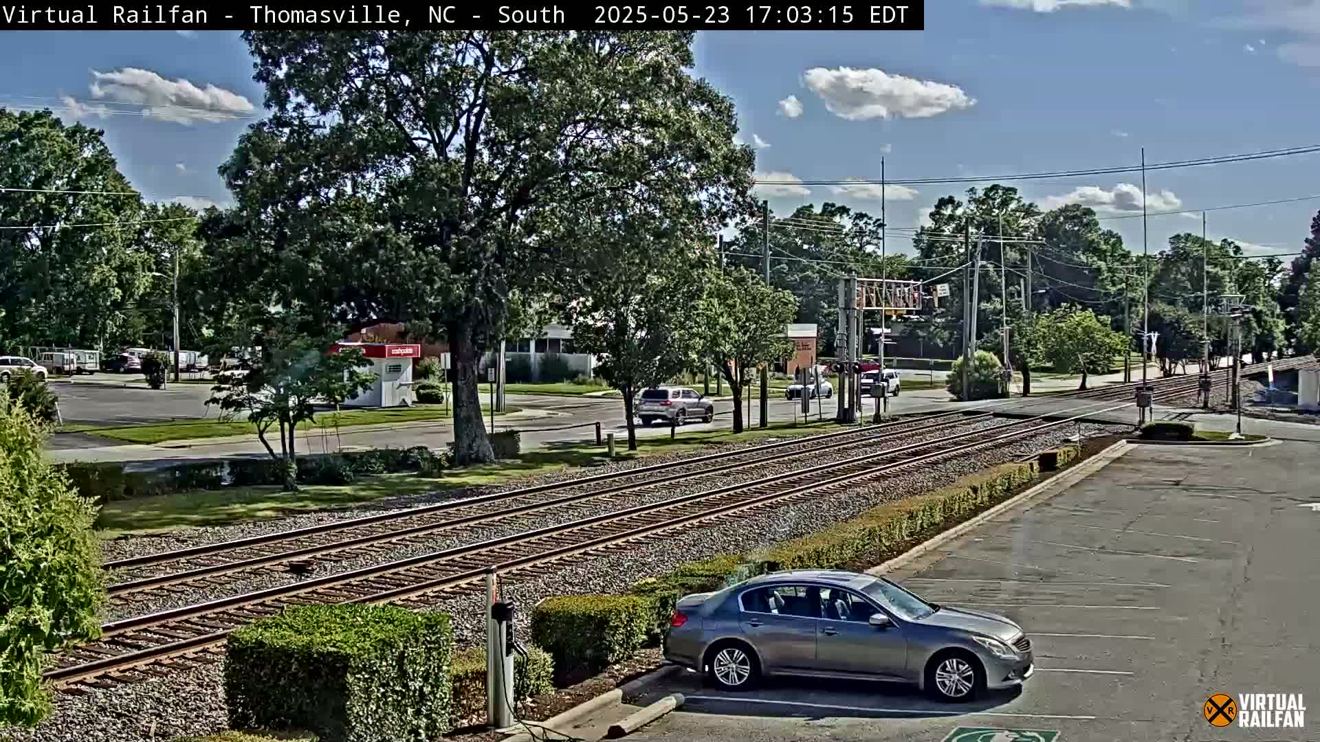 A partly sunny day shows train tracks intersecting a road with several cars visible, surrounded by lush green trees and shrubbery.