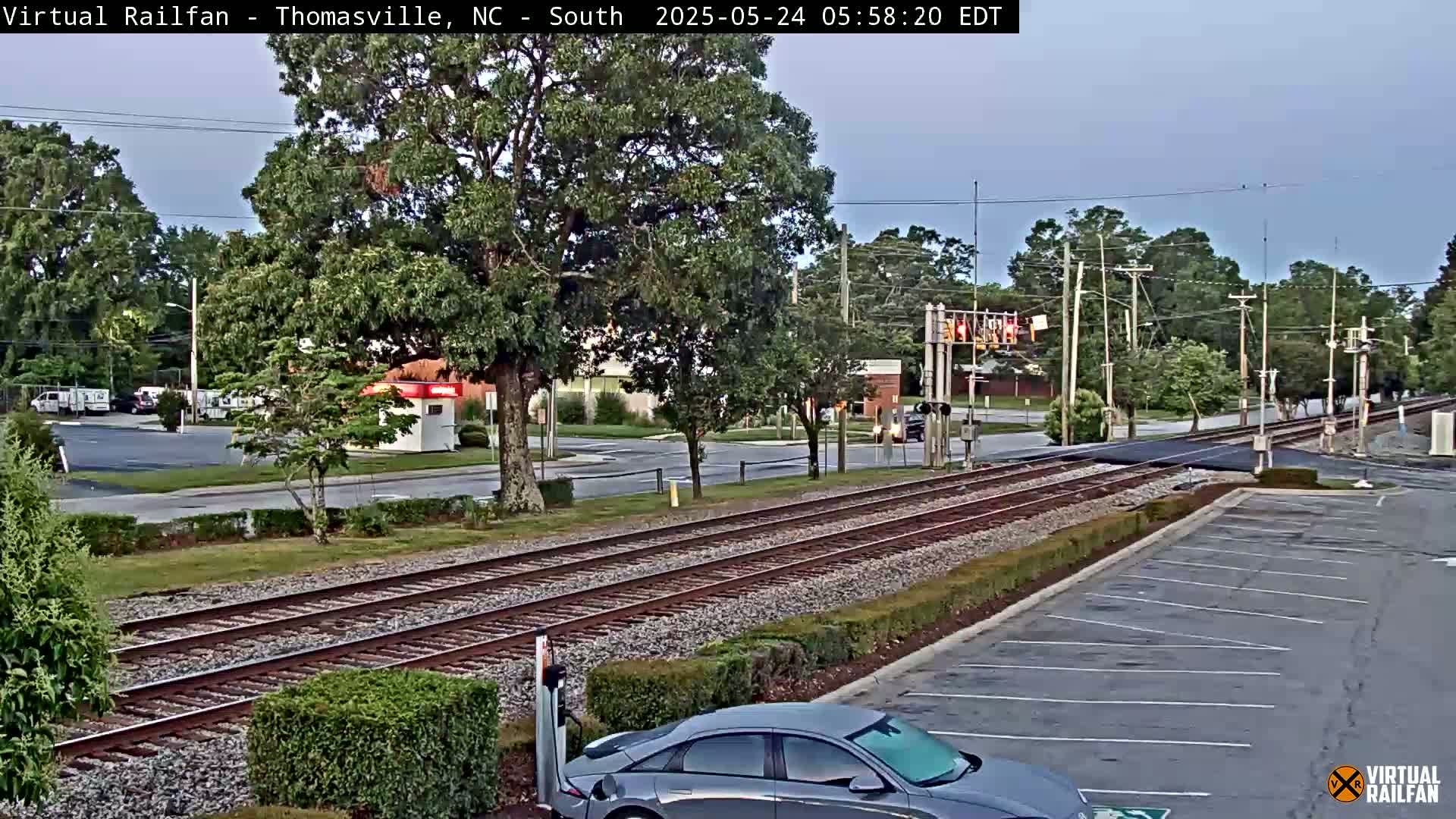 A view of railroad tracks crossing a road, under a mostly clear sky, with cars parked in a lot and vehicles driving on the road.