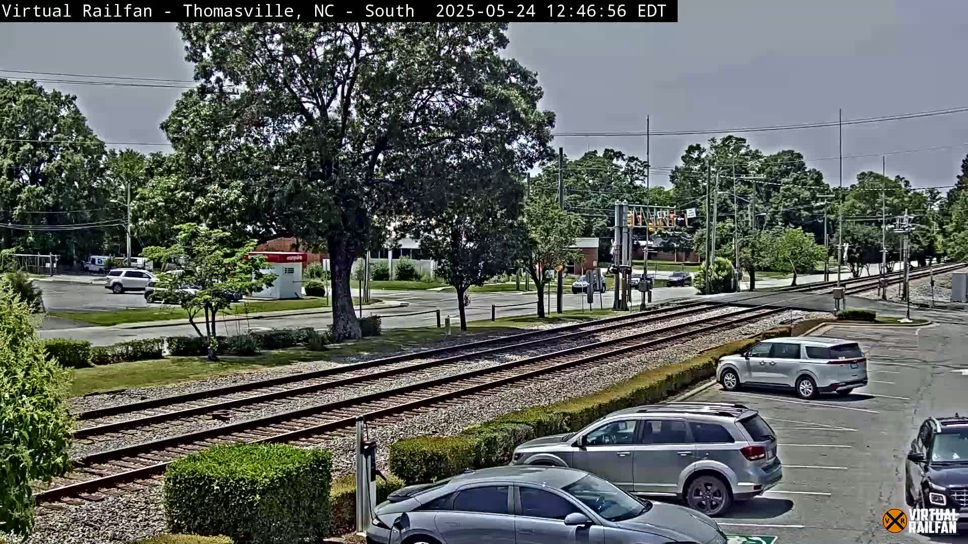 A mostly sunny outdoor scene shows railroad tracks intersecting a road, with several cars parked in a lot nearby, and leafy green trees lining the street.