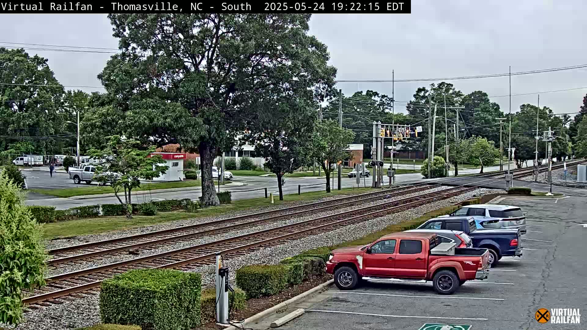 A parking lot with several vehicles, including a red pickup truck, is adjacent to railroad tracks that cross a road under an overcast sky.