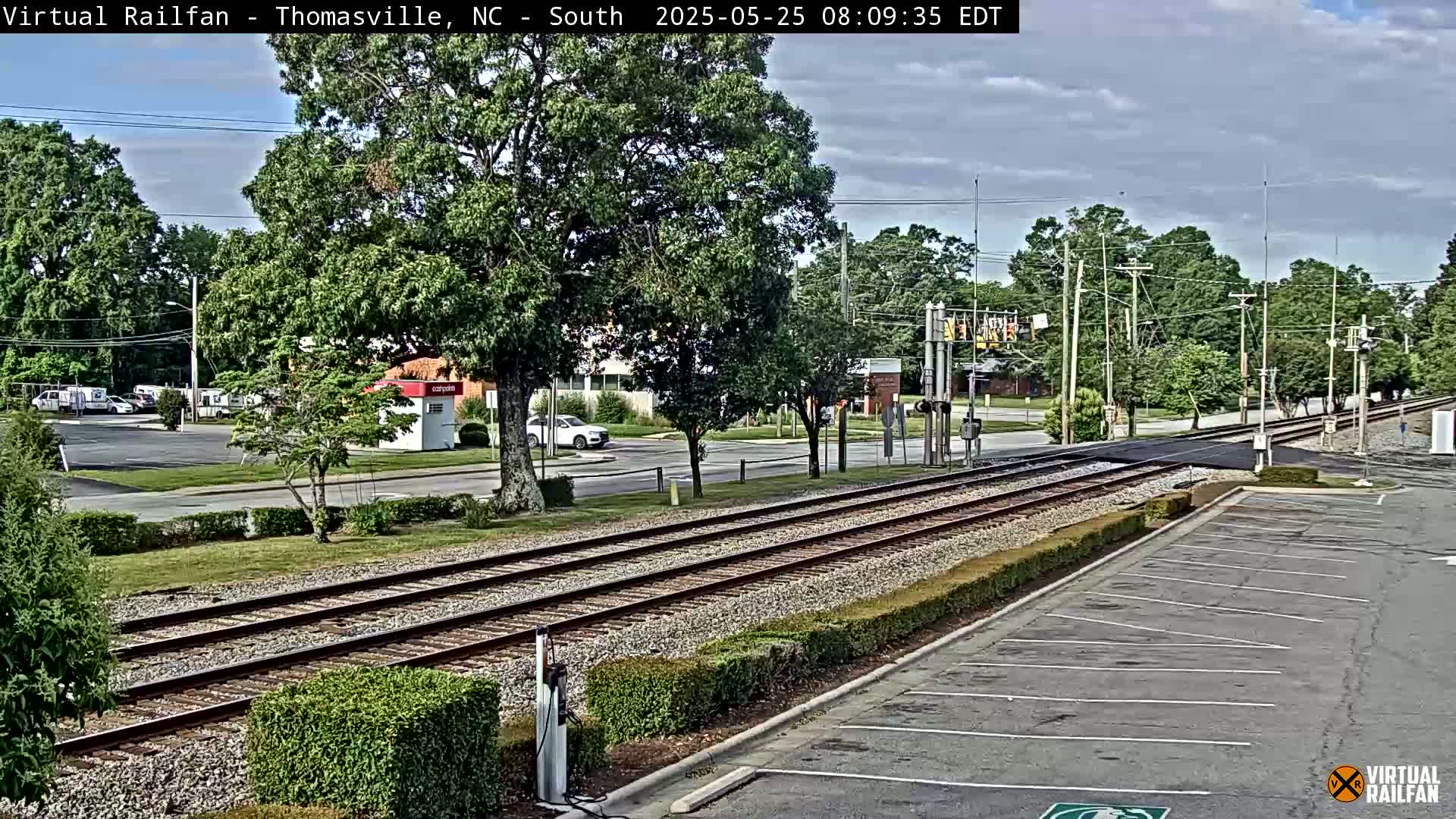 A mostly sunny day shows a view of train tracks that cross a road lined with trees and some buildings, with an empty parking lot next to the tracks.