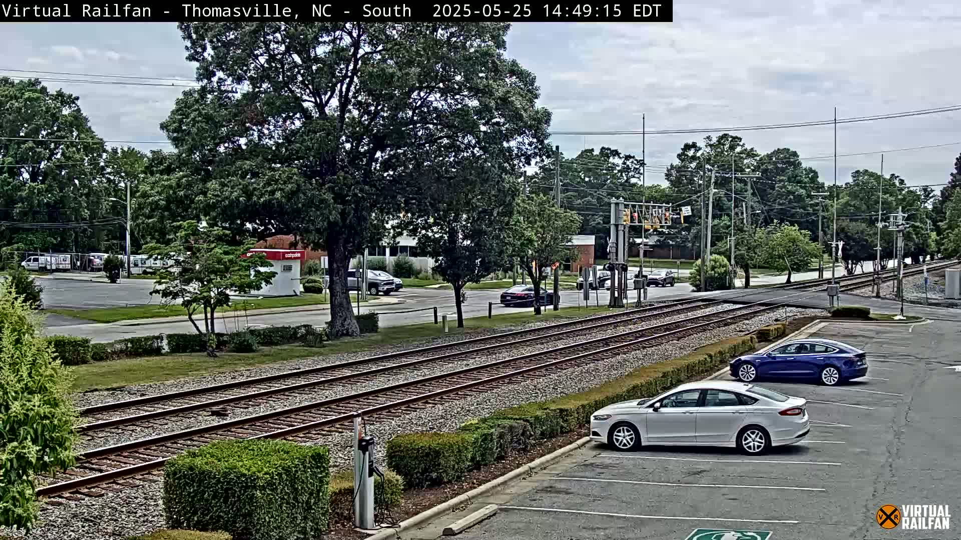 A view of a railroad crossing with several cars parked nearby, under partly cloudy skies.