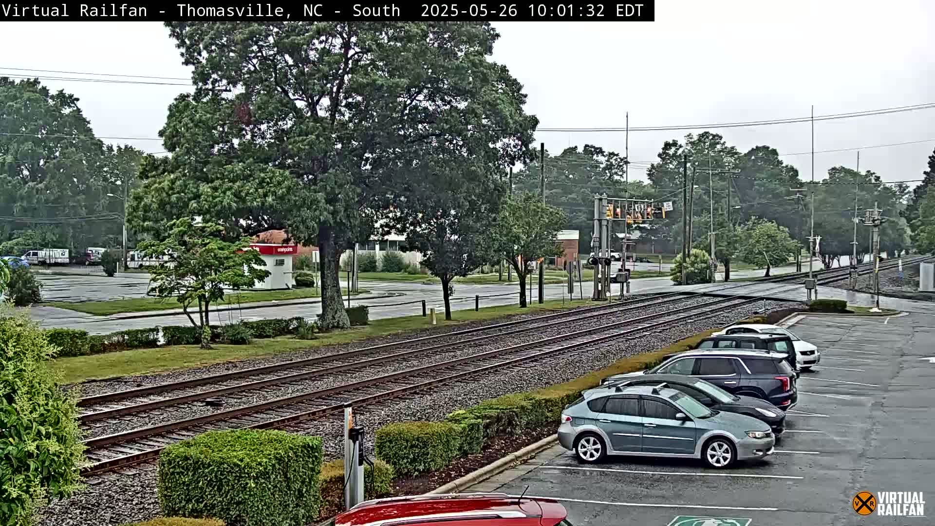 A view of railroad tracks crossing a street in a lightly wooded area on a rainy day, with several cars parked in a lot nearby.