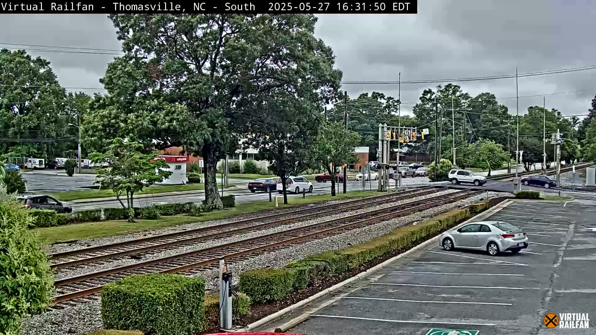 A view of a railroad crossing in a town on an overcast day, with several cars visible on the road and in a parking lot.