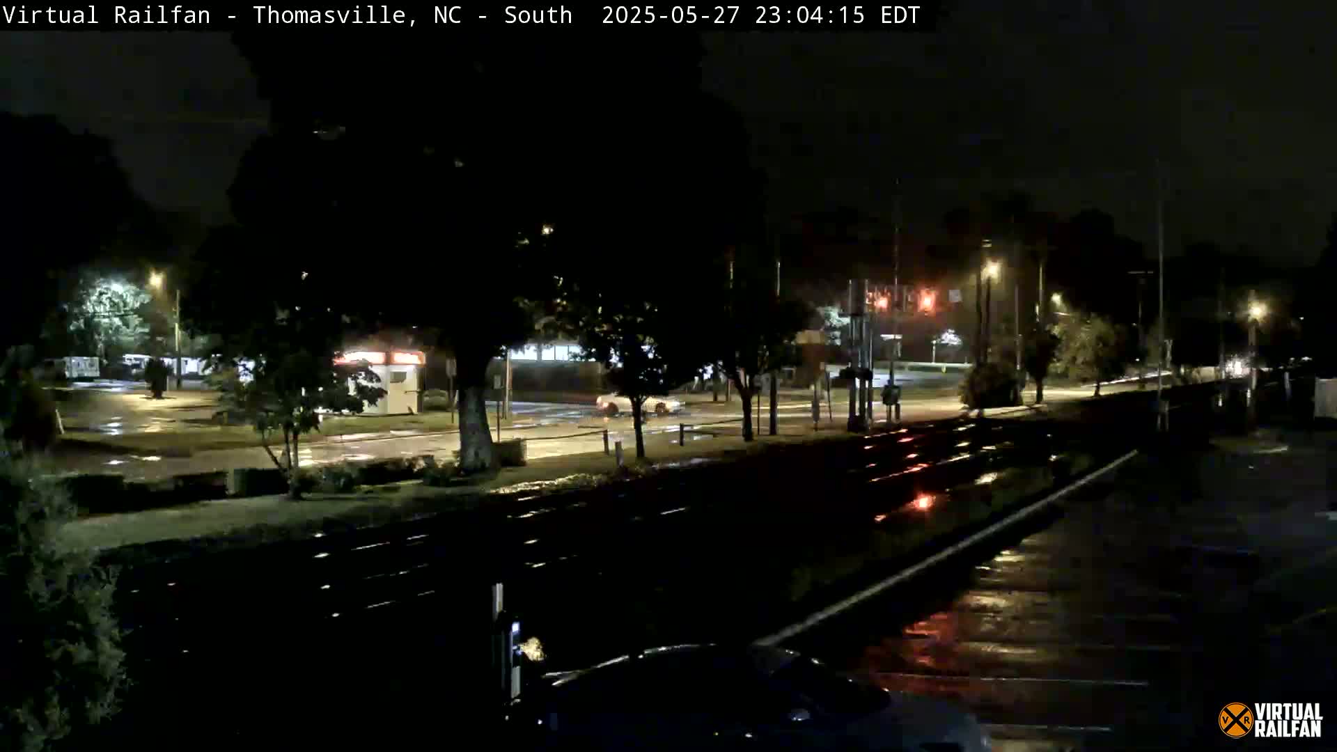 A nighttime view of rain-slicked streets and railroad tracks near a road intersection, with sparse traffic and streetlights illuminating the scene.