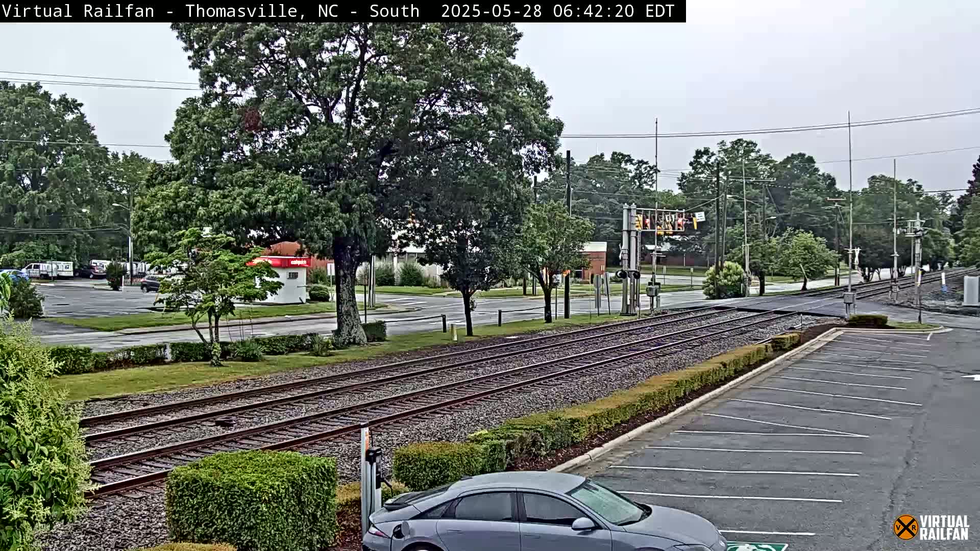 A view of railroad tracks crossing a street in an overcast setting, with a car parked in a lot and some buildings and trees in the background.