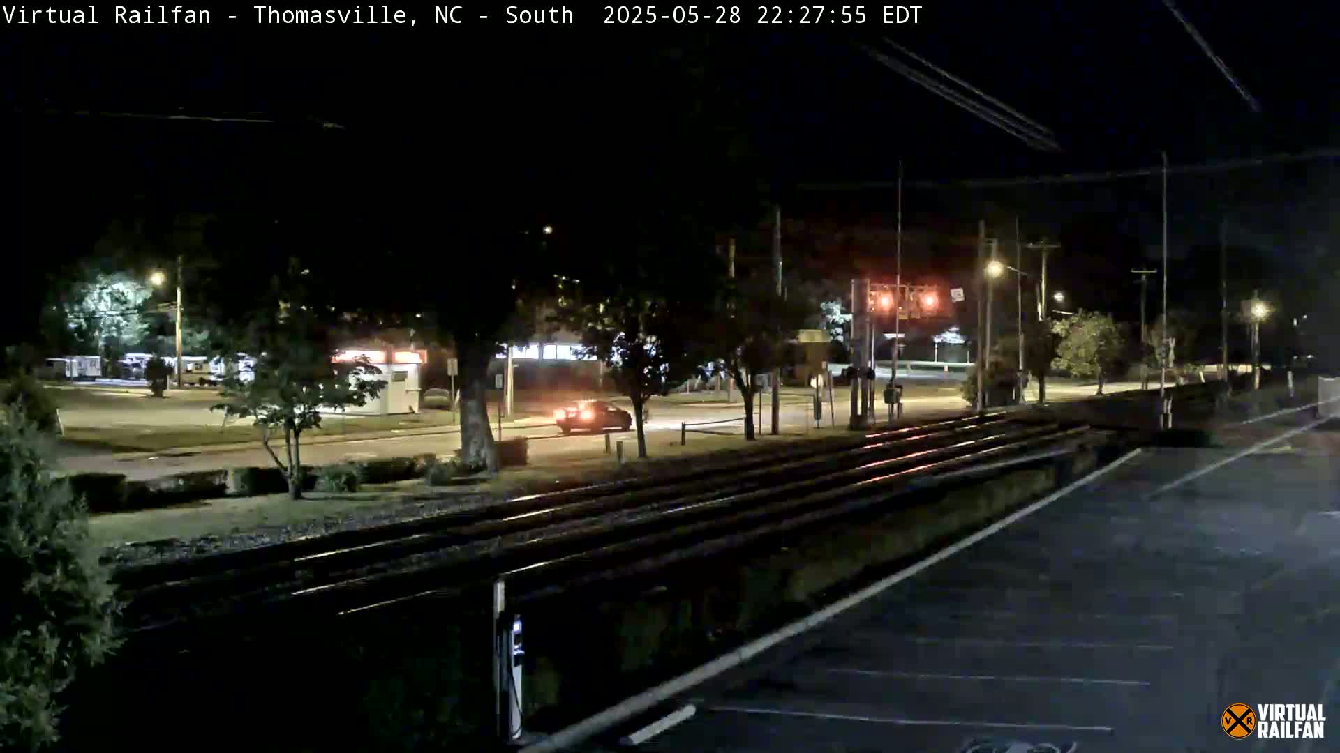 A nighttime view of a roadway and railroad tracks, with a single car visible on the road under clear skies.