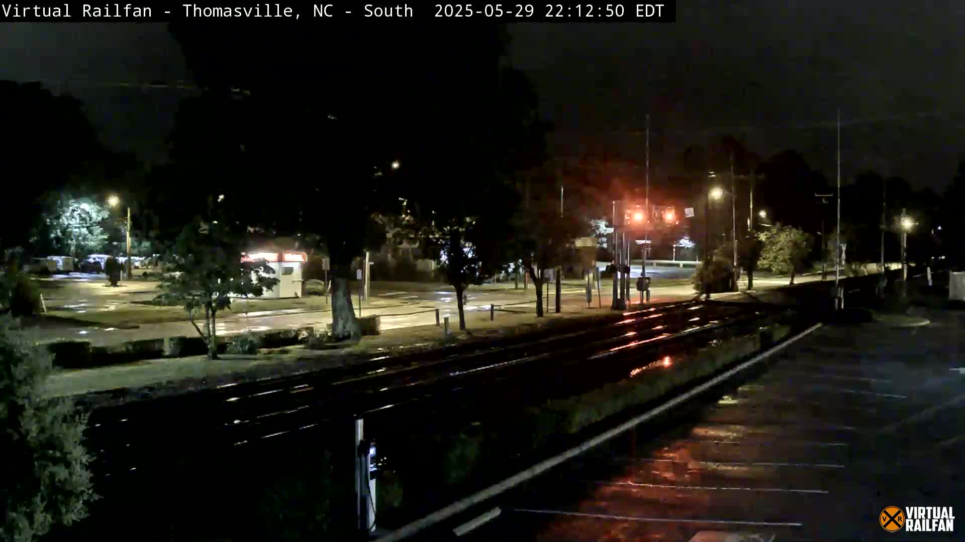 A nighttime view of a wet train track intersecting with a parking lot, under streetlights in the rain.
