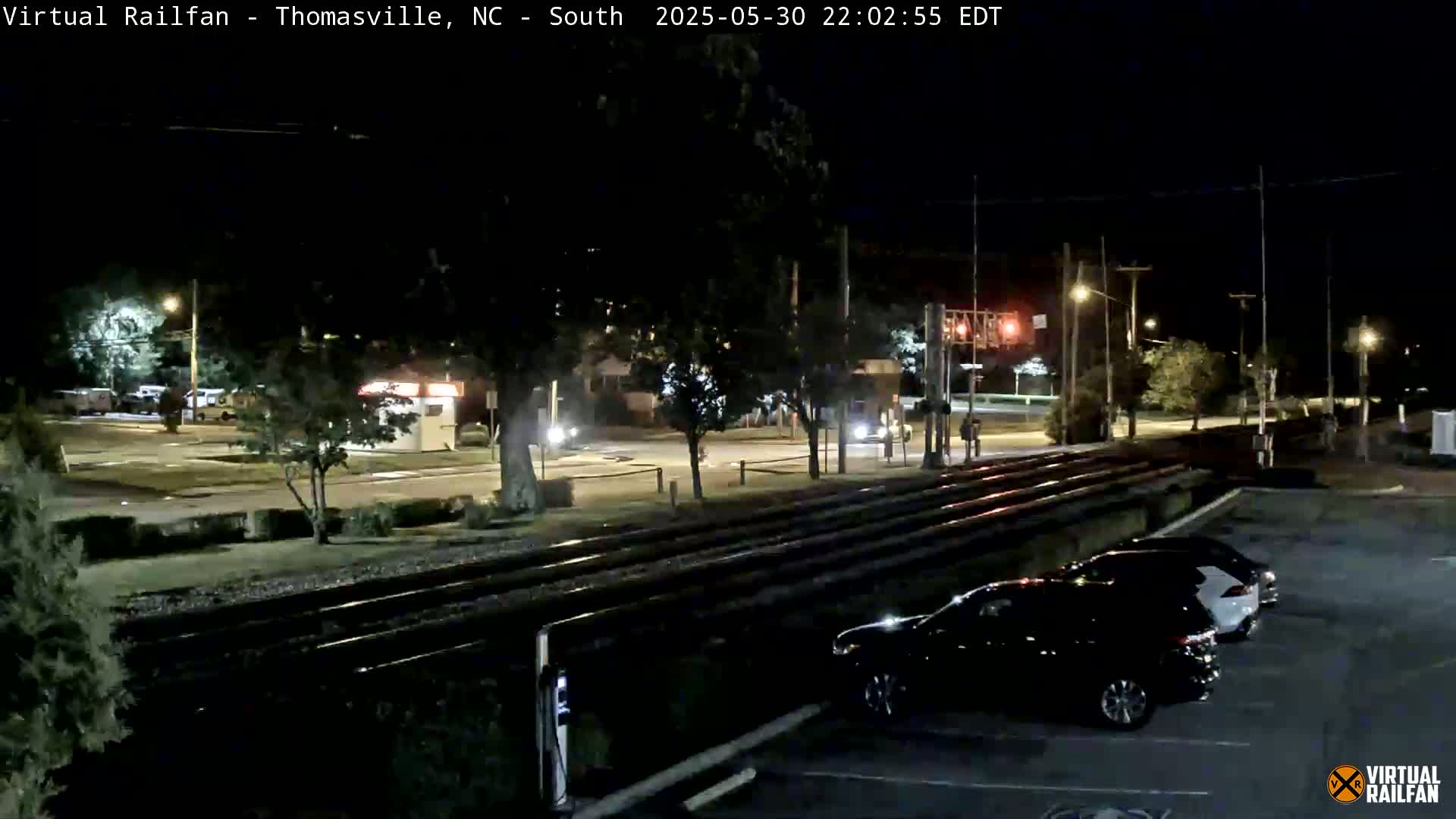 A nighttime view of a railroad crossing with cars parked nearby under a clear, dark sky.