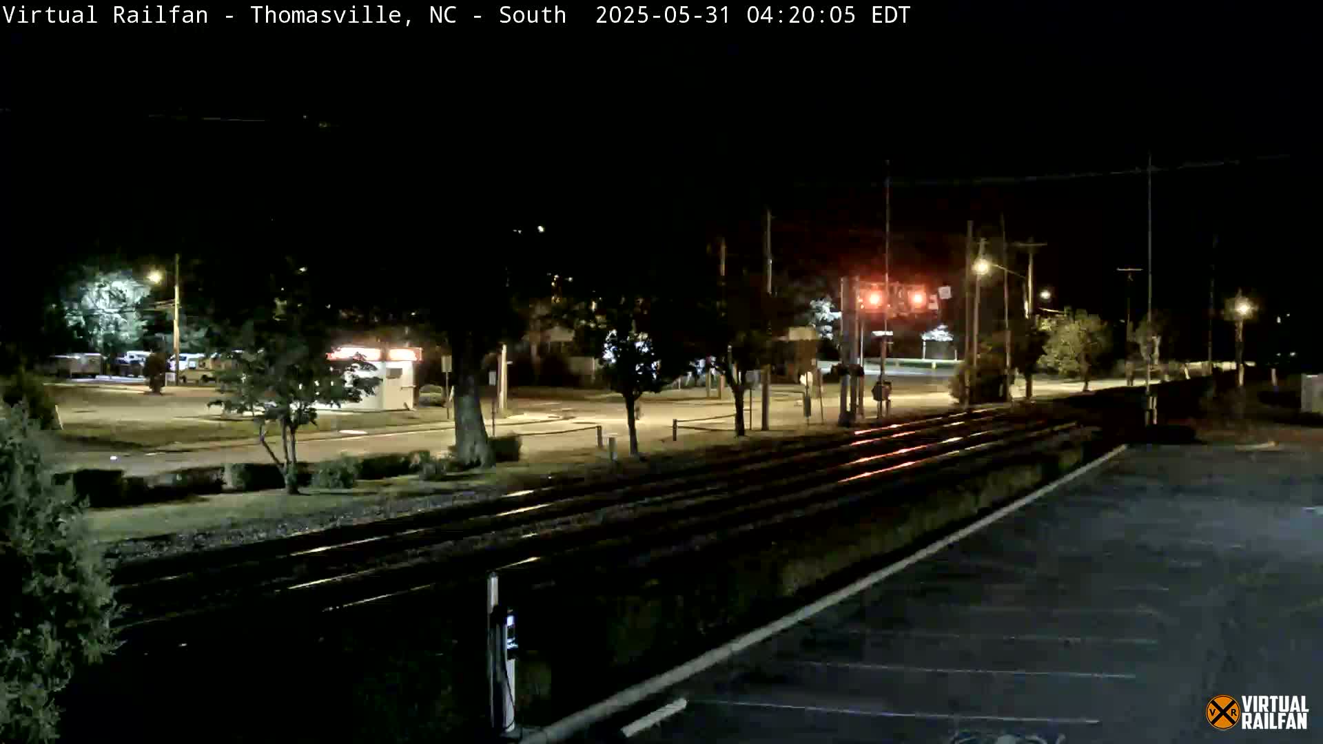 A nighttime view of railroad tracks next to a street, with a few cars parked nearby and trees lining the street.