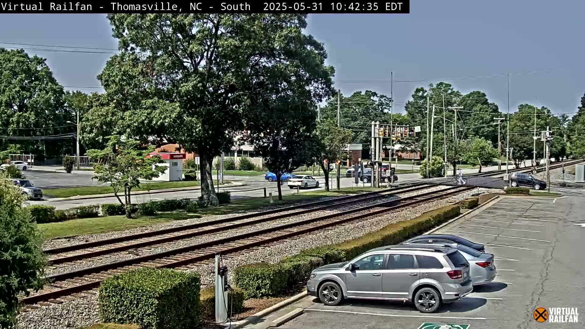 A sunny day view of railroad tracks crossing a road intersection, with several cars parked in a lot and other cars driving by.