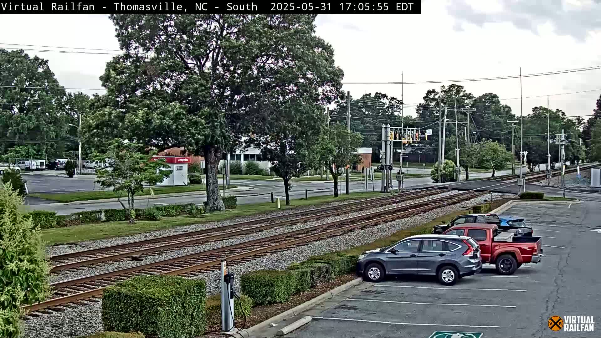 A view of railroad tracks running through a suburban area under a partly cloudy sky, with several cars parked nearby.