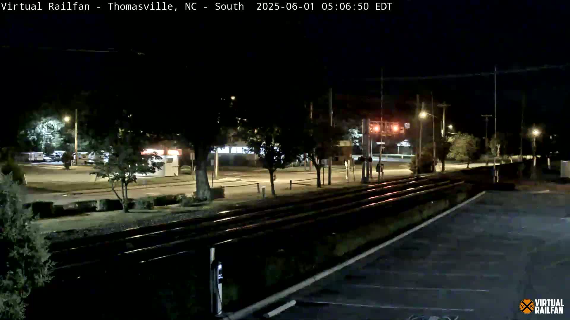 A nighttime view of train tracks next to a mostly empty parking lot and street, with a few streetlights illuminating the scene.