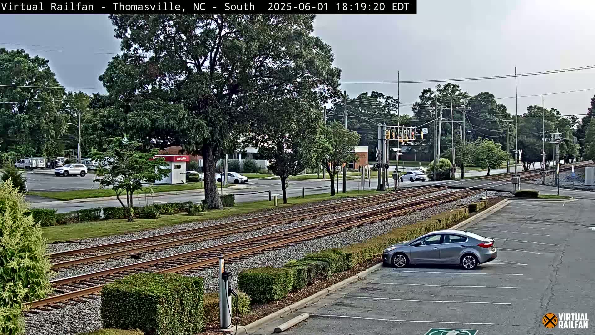 A train track runs through a suburban area under an overcast sky, with several cars parked nearby and a few driving on the road.