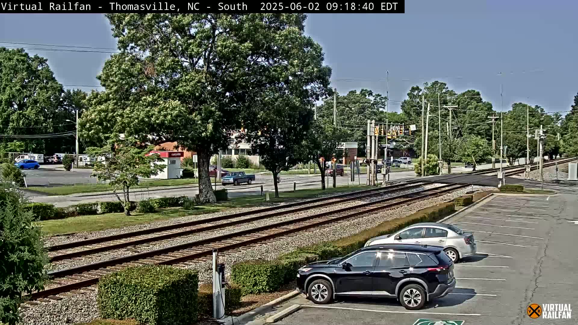 Under a clear sky, railroad tracks cross a road in a suburban area with several cars parked and driving near a small building.
