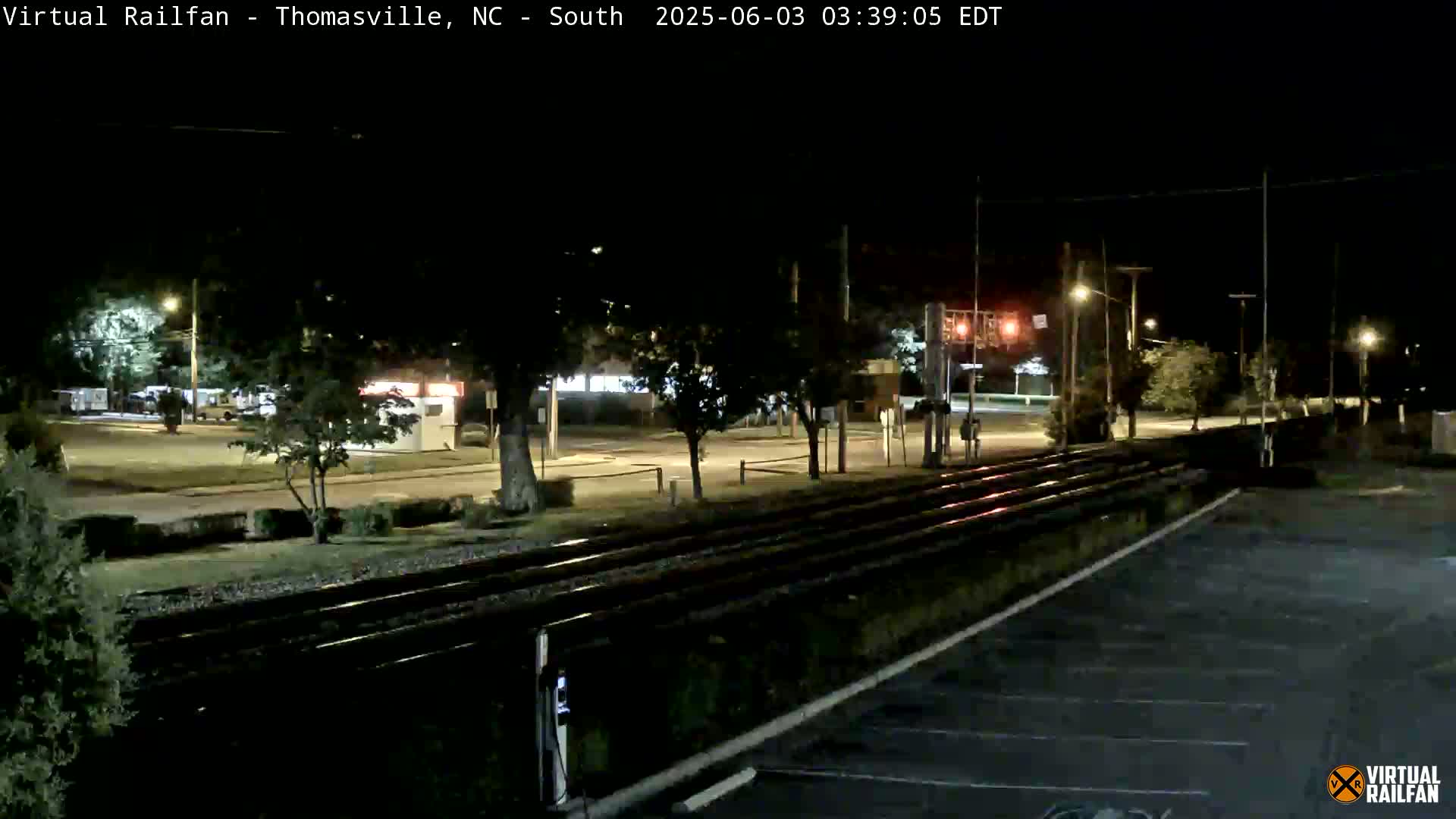 At night, a nighttime view shows train tracks next to a road with cars and streetlights, under a dark sky.