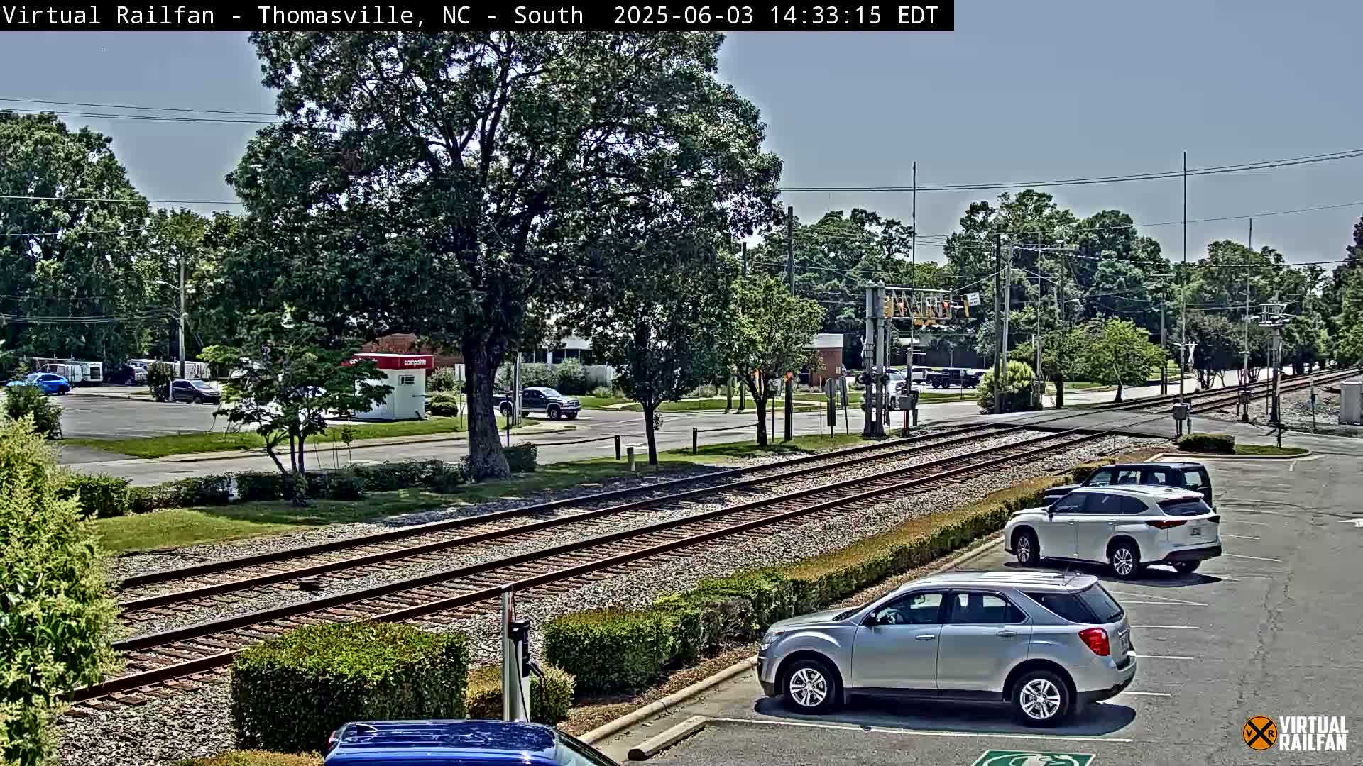 Under a mostly clear sky, railroad tracks run through a suburban area with several cars parked nearby.