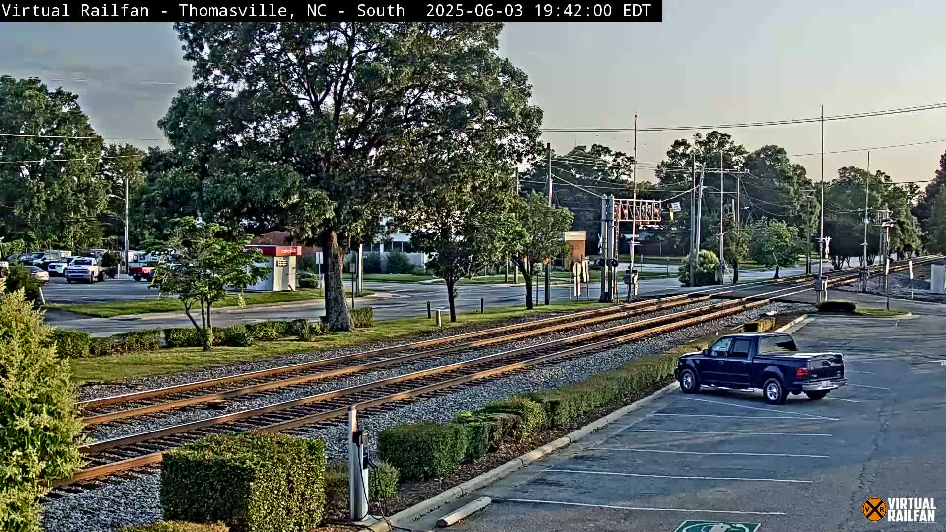 A dark-colored pickup truck is parked in a lot next to train tracks that run through a tree-lined area in the evening.