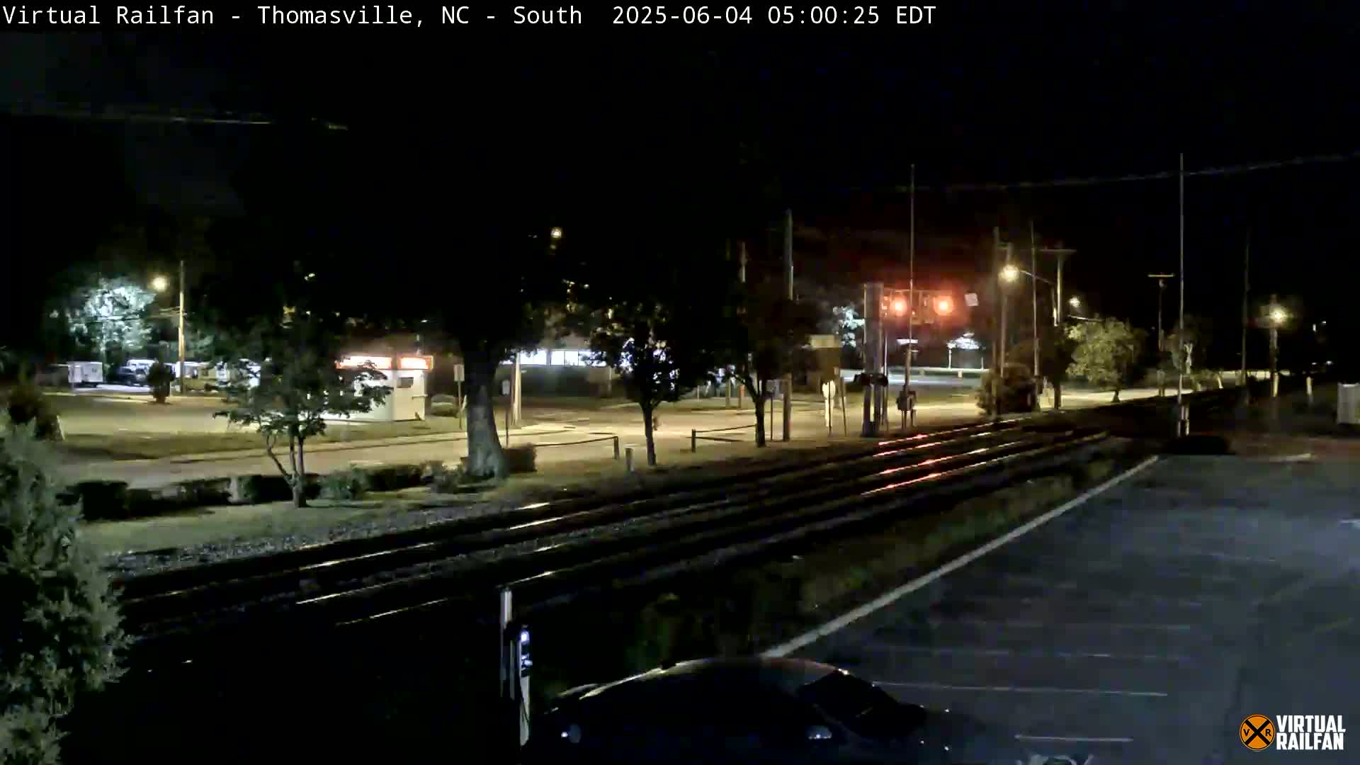 A nighttime view of railroad tracks intersecting a road, with streetlights illuminating sparsely populated surroundings.