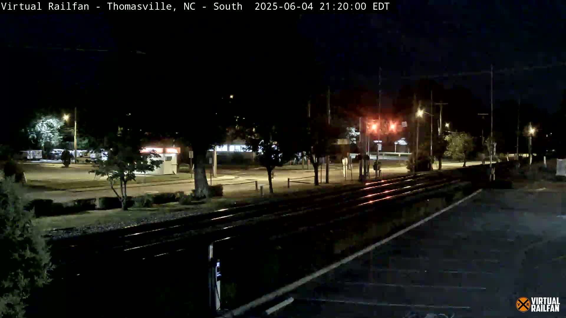 A nighttime view of train tracks running through a parking lot and alongside a road lined with trees and streetlights.