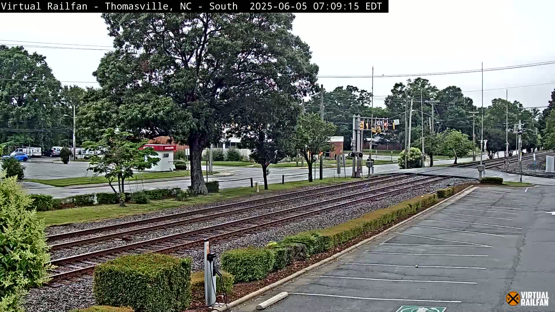 A view of railroad tracks crossing a road on an overcast day, with a parking lot and several buildings visible in the background.
